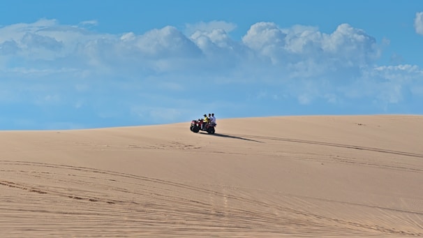 A person rides an ATV across expansive sand dunes under a vivid blue sky filled with scattered white clouds. The smooth sand has visible tire tracks, indicative of recent activity.