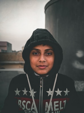 A young person is wearing a dark hooded jacket with a pattern of stars and text. The background is blurred with industrial structures giving an urban feel.