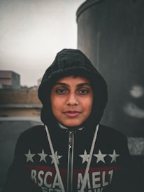 A young person is wearing a dark hooded jacket with a pattern of stars and text. The background is blurred with industrial structures giving an urban feel.
