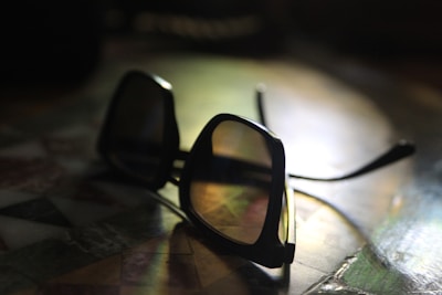 A pair of dark sunglasses with a glossy black frame rests on a colorful, tiled surface. The light casts subtle reflections on the lenses, highlighting the smooth curves of the frame.