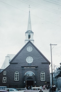A historical church building with a tall steeple and cross on top, featuring arched windows and a stone facade. The entrances are surrounded by people and several vintage cars are parked in front. The structure has a date inscribed on it and there are power lines visible in the background.