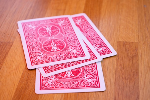 Several playing cards are spread on a wooden surface. The cards have a red and white intricate design on their backs.