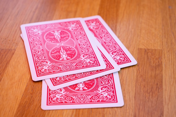 Several playing cards are spread on a wooden surface. The cards have a red and white intricate design on their backs.