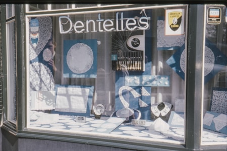 A shop window display features intricate lace patterns and doilies in white, set against a light blue backdrop. Several framed lace designs, as well as lace collars and other lace items, are arranged artistically. Signs indicate the acceptance of American Express cards, and the overall theme revolves around lace, highlighted by the word 'Dentelles' above the display.