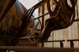 An industrial setting features rusty machinery, including a large metal cogwheel and various cylindrical and angular components. The equipment is surrounded by metal structures with a weathered, corroded surface, indicating age and disuse.