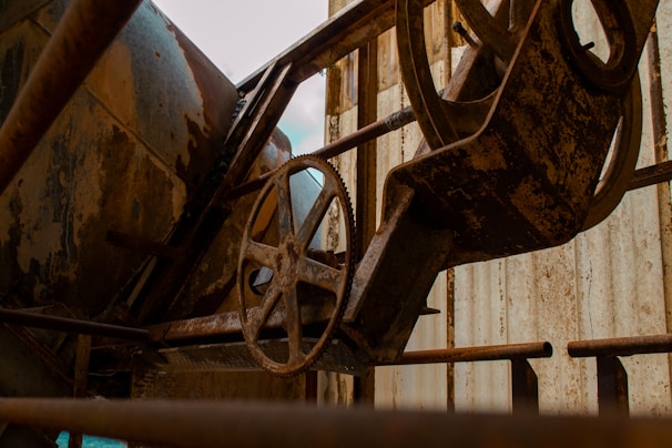An industrial setting features rusty machinery, including a large metal cogwheel and various cylindrical and angular components. The equipment is surrounded by metal structures with a weathered, corroded surface, indicating age and disuse.