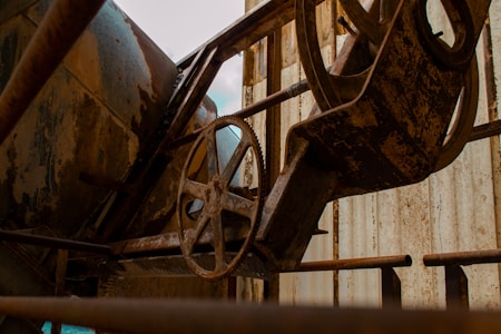 An industrial setting features rusty machinery, including a large metal cogwheel and various cylindrical and angular components. The equipment is surrounded by metal structures with a weathered, corroded surface, indicating age and disuse.