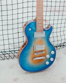 A blue electric guitar with a sunburst gradient leaning against a black netting. The guitar features chrome hardware, including two pickups, a bridge, and four control knobs. The setting appears to be outdoors on a white sandy surface.