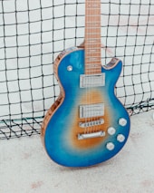 A blue electric guitar with a sunburst gradient leaning against a black netting. The guitar features chrome hardware, including two pickups, a bridge, and four control knobs. The setting appears to be outdoors on a white sandy surface.