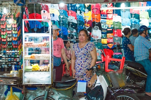 A bustling market scene featuring a stall filled with a variety of brightly colored shirts and footwear on display. A food cart with snacks and eggs occupies the foreground, beside which stands a woman in a floral dress. Several people are seen in the background, some browsing the merchandise.