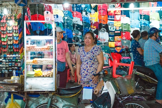 A bustling market scene featuring a stall filled with a variety of brightly colored shirts and footwear on display. A food cart with snacks and eggs occupies the foreground, beside which stands a woman in a floral dress. Several people are seen in the background, some browsing the merchandise.