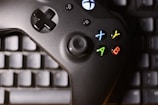 A black gaming controller with colored buttons, including X, Y, A, and B, is placed on top of a computer keyboard. The buttons are red, blue, yellow, and green, adding contrast to the dark controller and keyboard beneath.