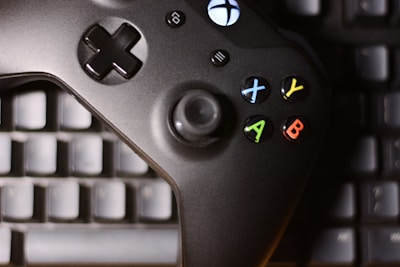 A black gaming controller with colored buttons, including X, Y, A, and B, is placed on top of a computer keyboard. The buttons are red, blue, yellow, and green, adding contrast to the dark controller and keyboard beneath.