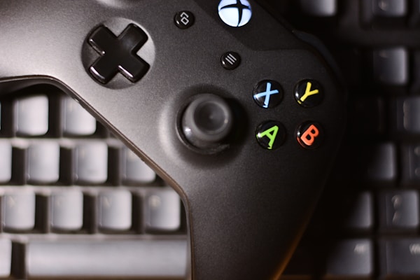 A black gaming controller with colored buttons, including X, Y, A, and B, is placed on top of a computer keyboard. The buttons are red, blue, yellow, and green, adding contrast to the dark controller and keyboard beneath.