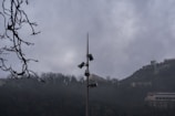 A tall pole with multiple security cameras and loudspeakers is situated in the middle of a misty, mountainous area. Bare tree branches are visible in the foreground, while buildings and infrastructure are seen on the hills in the background. The sky is overcast, contributing to a gloomy atmosphere.