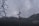 A tall pole with multiple security cameras and loudspeakers is situated in the middle of a misty, mountainous area. Bare tree branches are visible in the foreground, while buildings and infrastructure are seen on the hills in the background. The sky is overcast, contributing to a gloomy atmosphere.