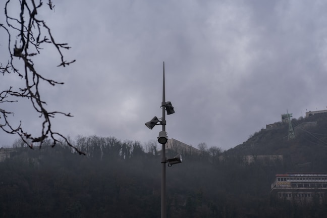 A tall pole with multiple security cameras and loudspeakers is situated in the middle of a misty, mountainous area. Bare tree branches are visible in the foreground, while buildings and infrastructure are seen on the hills in the background. The sky is overcast, contributing to a gloomy atmosphere.