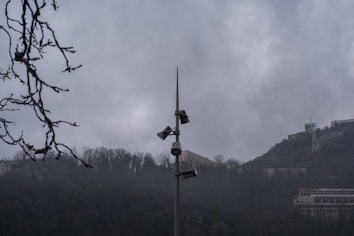 A tall pole with multiple security cameras and loudspeakers is situated in the middle of a misty, mountainous area. Bare tree branches are visible in the foreground, while buildings and infrastructure are seen on the hills in the background. The sky is overcast, contributing to a gloomy atmosphere.