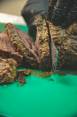 A person wearing black gloves slices brisket on a green cutting board. The meat appears juicy and well-cooked, with a browned crust and tender interior. The knife is in mid-cut, indicating the meat is being prepared for serving.
