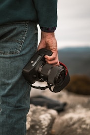 A close-up view of a person wearing blue jeans and a dark jacket, holding a professional camera by their side. The background is a blurred natural landscape with rocky surfaces, suggesting an outdoor setting.