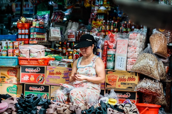 A vendor sits in a bustling market stall surrounded by various packaged food products, including instant noodles, snacks, and canned goods. The vendor, wearing a floral dress and a black cap, is engaged with her hands, possibly sorting or packaging items. The space is filled with colorful and neatly arranged products in boxes and bags, creating a vibrant and busy atmosphere.