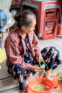 An elderly woman with graying hair tied up is sitting on the steps, peeling or cleaning green vegetables. She is wearing a red plaid shirt over a floral patterned dress. In front of her, there is a red plastic basket containing more green vegetables. Behind her, there is a stack of red plastic stools.