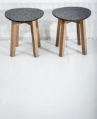 Two wooden stools with dark gray circular seats rest on a light-colored floor. The stools have three legs made of light wood, providing a simple and minimalist design.
