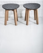 Two wooden stools with dark gray circular seats rest on a light-colored floor. The stools have three legs made of light wood, providing a simple and minimalist design.