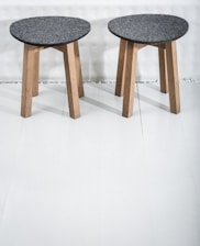 Two wooden stools with dark gray circular seats rest on a light-colored floor. The stools have three legs made of light wood, providing a simple and minimalist design.