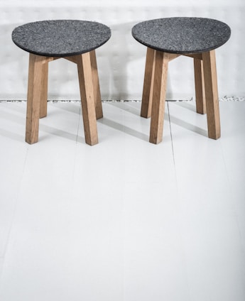 Two wooden stools with dark gray circular seats rest on a light-colored floor. The stools have three legs made of light wood, providing a simple and minimalist design.