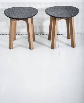 Two wooden stools with dark gray circular seats rest on a light-colored floor. The stools have three legs made of light wood, providing a simple and minimalist design.