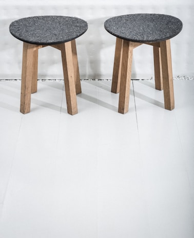 Two wooden stools with dark gray circular seats rest on a light-colored floor. The stools have three legs made of light wood, providing a simple and minimalist design.