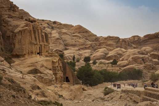 A rugged landscape featuring ancient rock-cut architecture in a desert environment. A large cliff face dominates the left side with carved structures, while a pathway on the right shows small groups of people walking and a horse-drawn carriage. Sparse greenery is scattered among the rocky terrain under a subtly overcast sky.