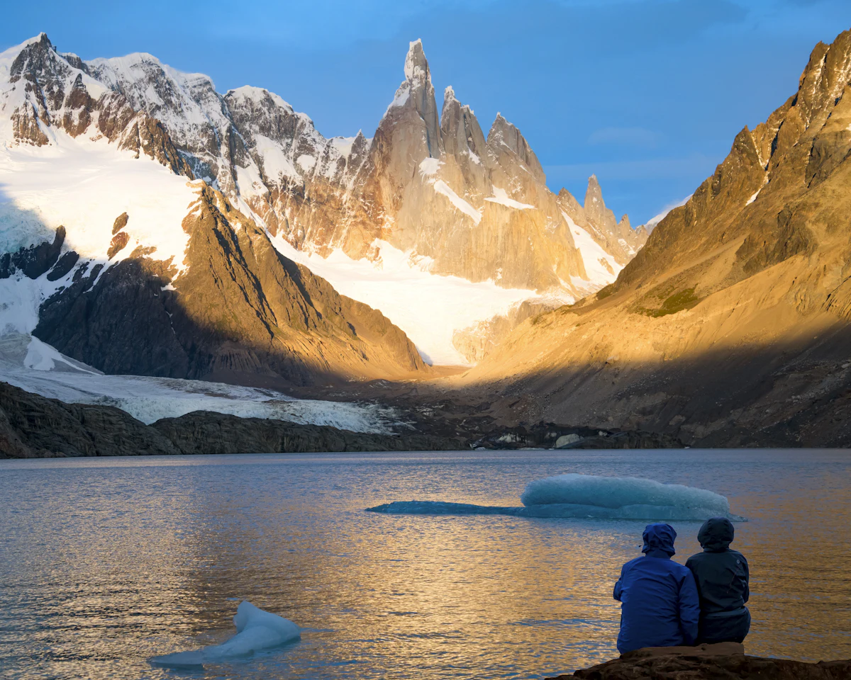 Two hikers sit on the shore of Laguna Torre, with icebergs in the lake and Cerro Torre glowing gold at sunrise above the glacier.