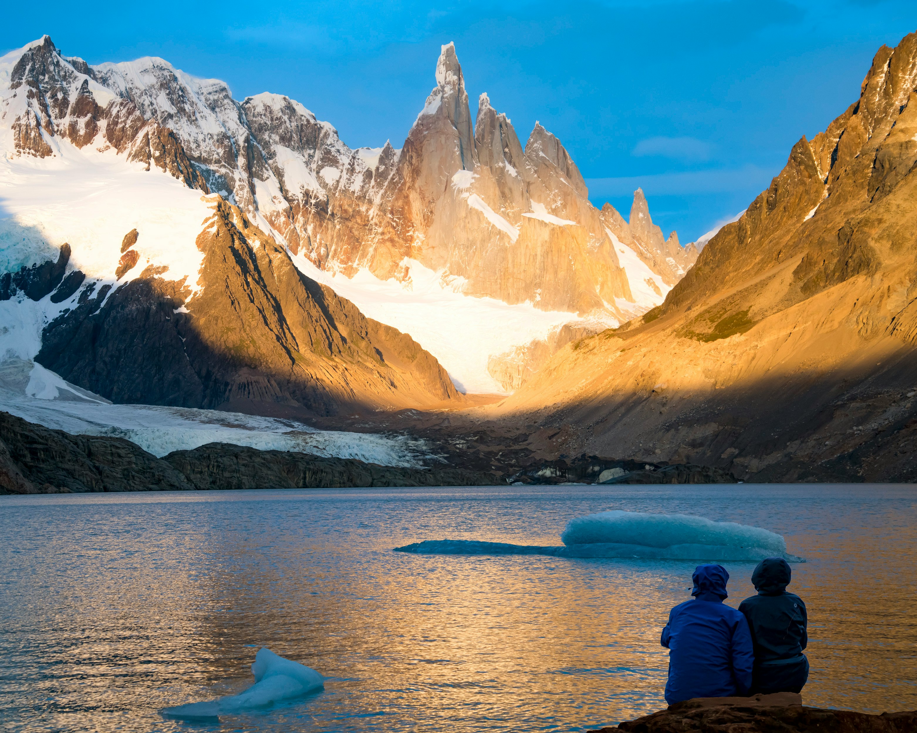 person in black jacket sitting on rock in front of lake and snow covered mountain