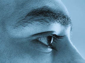 A close-up view of the side of a human face, focusing on the eye and eyebrow. The image has a blue tint, highlighting the eyelashes and texture of the skin.