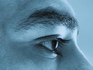 A close-up view of the side of a human face, focusing on the eye and eyebrow. The image has a blue tint, highlighting the eyelashes and texture of the skin.