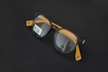 Round black eyeglasses with orange arms are placed on a dark surface. A barcode sticker is visible on one of the lenses, and the Converse logo is printed on the inside of the lenses.
