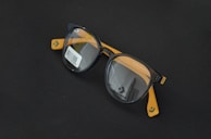 Round black eyeglasses with orange arms are placed on a dark surface. A barcode sticker is visible on one of the lenses, and the Converse logo is printed on the inside of the lenses.