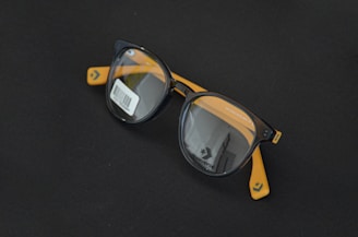 Round black eyeglasses with orange arms are placed on a dark surface. A barcode sticker is visible on one of the lenses, and the Converse logo is printed on the inside of the lenses.