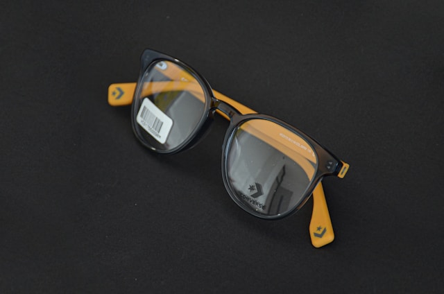 Round black eyeglasses with orange arms are placed on a dark surface. A barcode sticker is visible on one of the lenses, and the Converse logo is printed on the inside of the lenses.