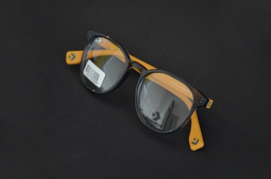 Round black eyeglasses with orange arms are placed on a dark surface. A barcode sticker is visible on one of the lenses, and the Converse logo is printed on the inside of the lenses.