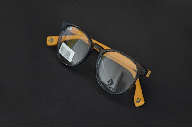 Round black eyeglasses with orange arms are placed on a dark surface. A barcode sticker is visible on one of the lenses, and the Converse logo is printed on the inside of the lenses.