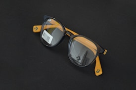 Round black eyeglasses with orange arms are placed on a dark surface. A barcode sticker is visible on one of the lenses, and the Converse logo is printed on the inside of the lenses.