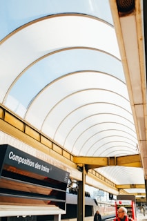 A modern train station features an arched roof with translucent panels allowing natural light. A digital sign displays 'Composition des trains' and several passengers can be seen near a train. A vending machine and a timetable board are visible.