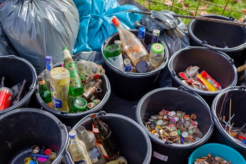 Several buckets are filled with various discarded items such as empty glass bottles, plastic bottle caps, and metal lids. Surrounding the buckets are large garbage bags in shades of grey and blue. Some buckets contain mixed debris including cans and wrappers.