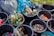 Several buckets are filled with various discarded items such as empty glass bottles, plastic bottle caps, and metal lids. Surrounding the buckets are large garbage bags in shades of grey and blue. Some buckets contain mixed debris including cans and wrappers.