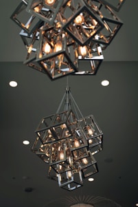 A modern chandelier composed of geometric metal frames encasing several glowing light bulbs, hanging from the ceiling. The ceiling background features several recessed lights.