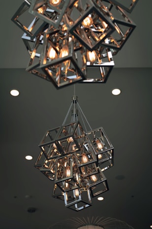 A modern chandelier composed of geometric metal frames encasing several glowing light bulbs, hanging from the ceiling. The ceiling background features several recessed lights.