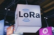 A brightly lit cube with the text 'LoRa' hangs from a ceiling, surrounded by various other illuminated signage. Industrial framework and lighting are visible in the background, suggesting a trade show or exhibition setting.